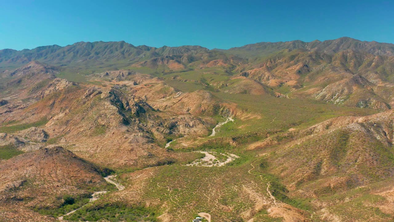 Wide aerial frame over Cabo Pulmo, Baja California Sur, showing brown desert hills, green valley vegetation, pale winding roads, distant ridges, clear blue sky, and no visible people.