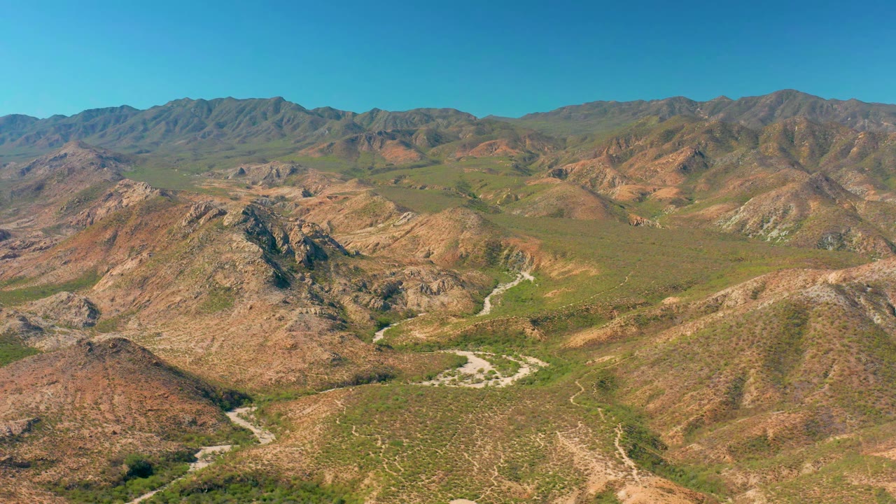 Aerial view in Cabo Pulmo, Baja California Sur, with the winding road more centered between brown slopes, green scrub patches, distant mountain ridges, blue sky, and no visible people.