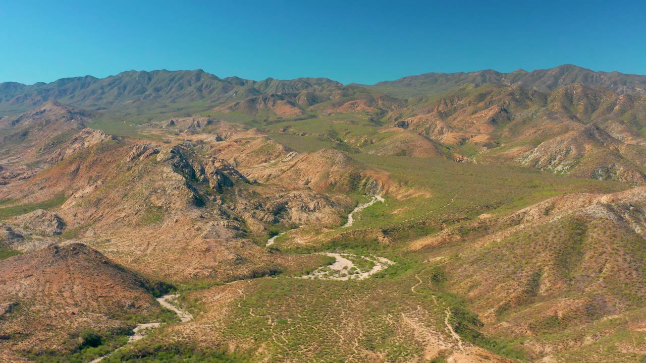 Mid-clip aerial frame in Cabo Pulmo, Baja California Sur, with rocky hills filling the lower frame, the winding dirt road visible through the valley, distant brown ridges, clear sky, and no visible people.