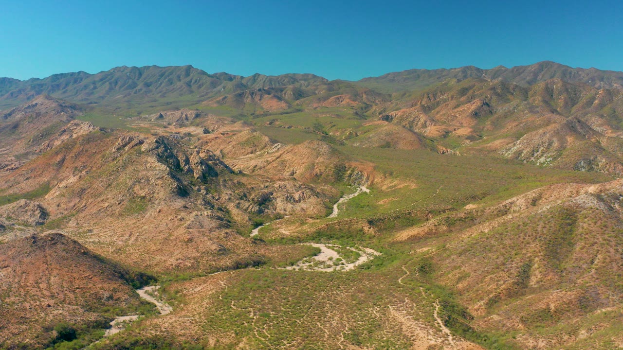 Opening aerial view over Cabo Pulmo hills with a pale winding road crossing the valley, distant ridges on the horizon, and no visible people.