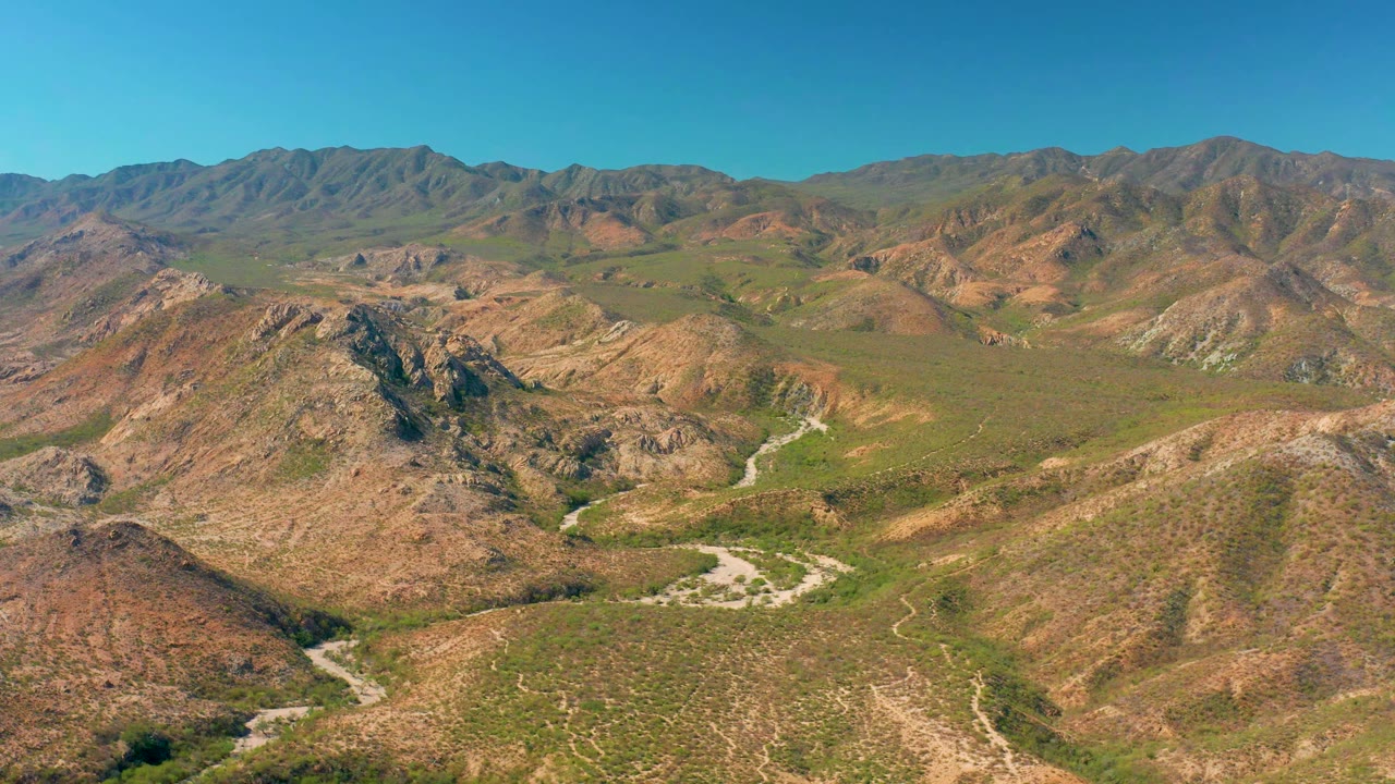 Aerial frame over Cabo Pulmo showing green-brown desert hills, a curving road near the lower valley, distant mountains, and no visible people.