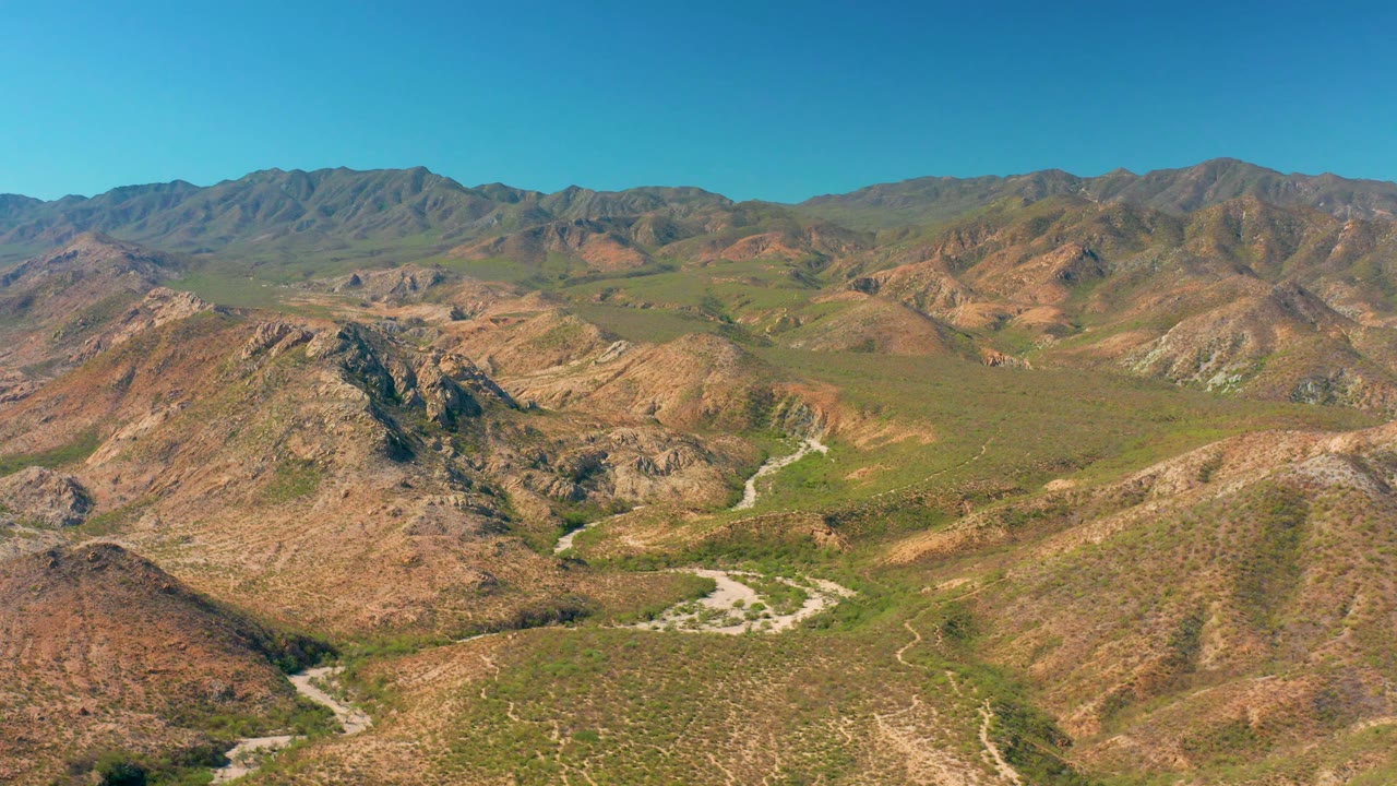Cabo Pulmo mountain valley seen from above, with dry ridges, scattered green vegetation, a winding road near the lower center, and no visible people.