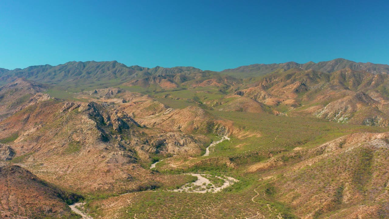 Opening aerial frame over Cabo Pulmo, Baja California Sur, with rocky brown hills, green scrub, a pale winding dirt road, distant mountains, and no visible people.