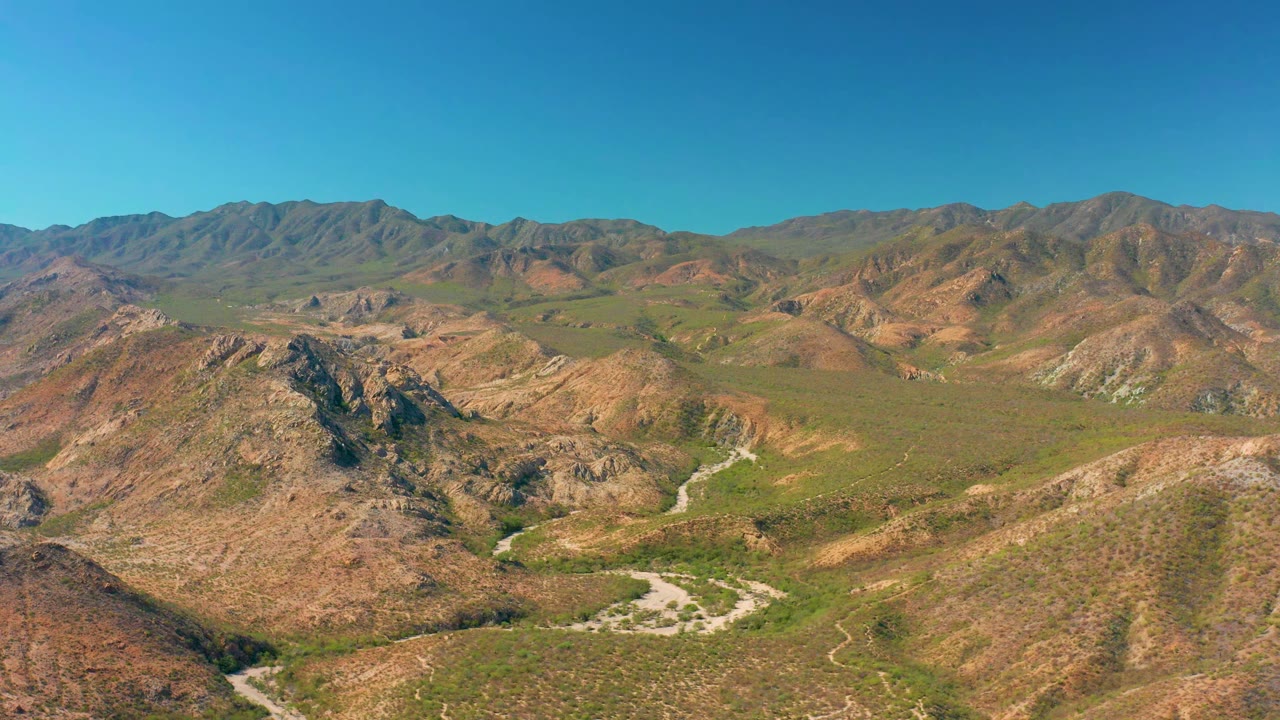 A wide aerial view in Cabo Pulmo shows the dirt road threading through green and brown valley terrain between rocky ridges, with no visible people.