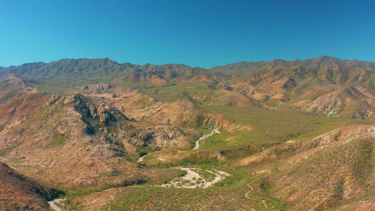 Opening aerial frame over Cabo Pulmo, Baja California Sur, with rocky green-brown hills, a pale road in the valley, distant mountains, and no visible people.