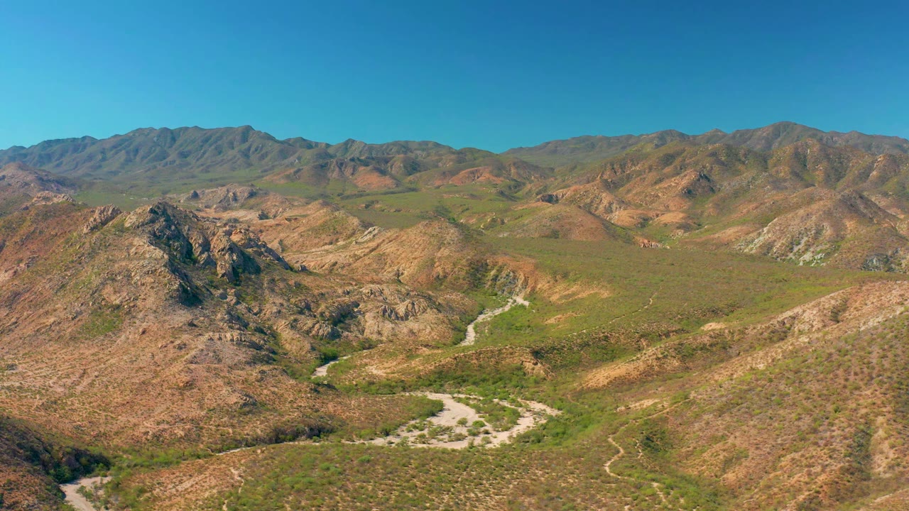 Cabo Pulmo aerial view with the winding pale road more centered between rocky slopes, scattered green vegetation, layered background mountains, and no visible people.