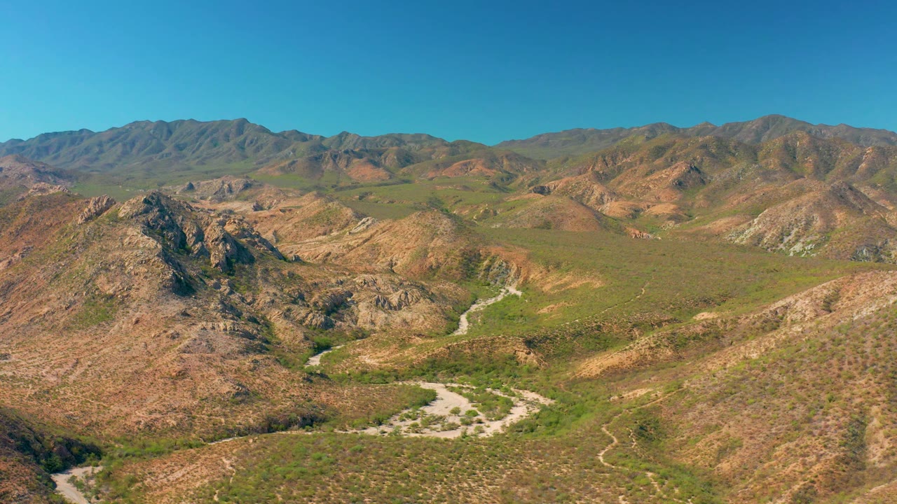 Aerial frame in Cabo Pulmo showing a rocky foreground hill, a pale road bending through green-brown scrubland, distant ridges across the horizon, and no visible people.