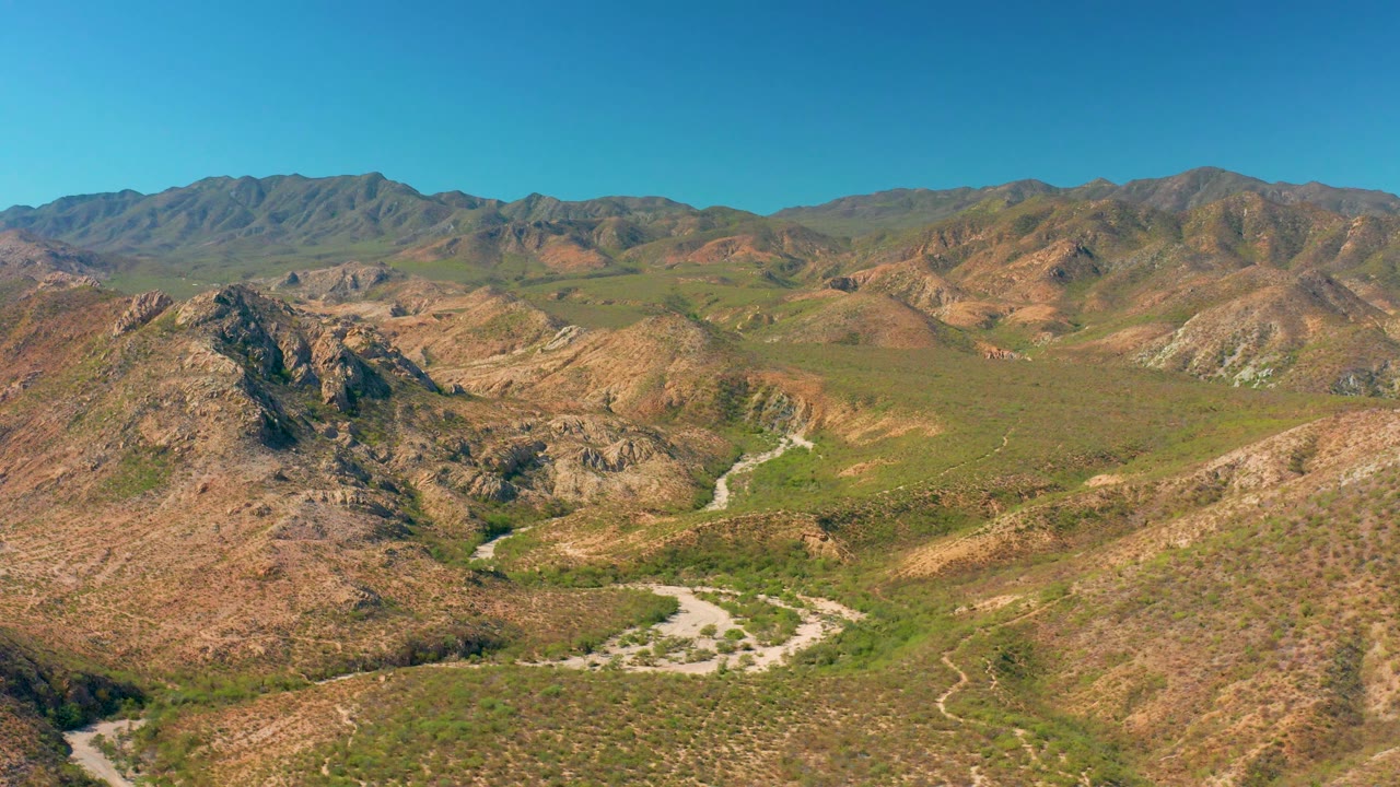 Cabo Pulmo mountain landscape with sunlit brown ridges, green vegetation patches across the valley, a winding road below, distant mountains, and no visible people.