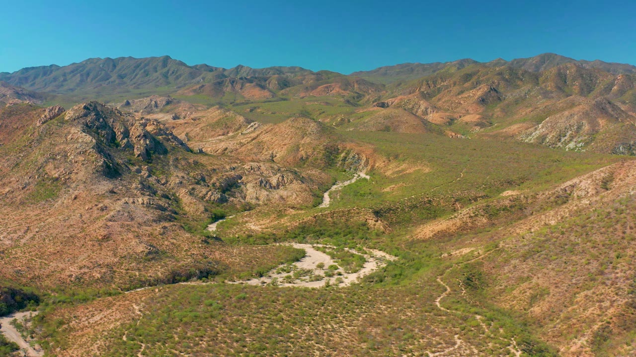 Aerial frame in Cabo Pulmo showing the dirt road lower in the valley, rocky slopes at left and right, layered mountain ridges in back, clear sky, and no visible people.