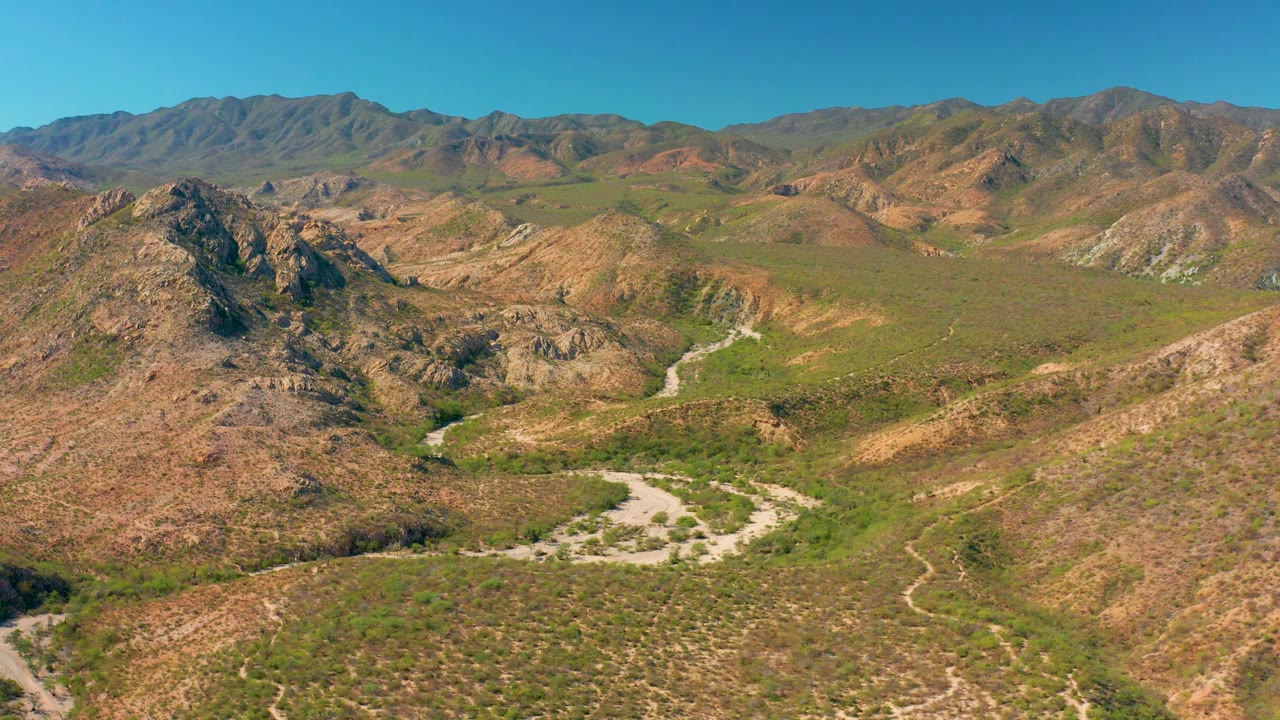 Mid-clip aerial frame over Cabo Pulmo terrain, with a jagged hill left of center, a winding dirt track below, broad scrubland, far ridgelines, and no visible people.