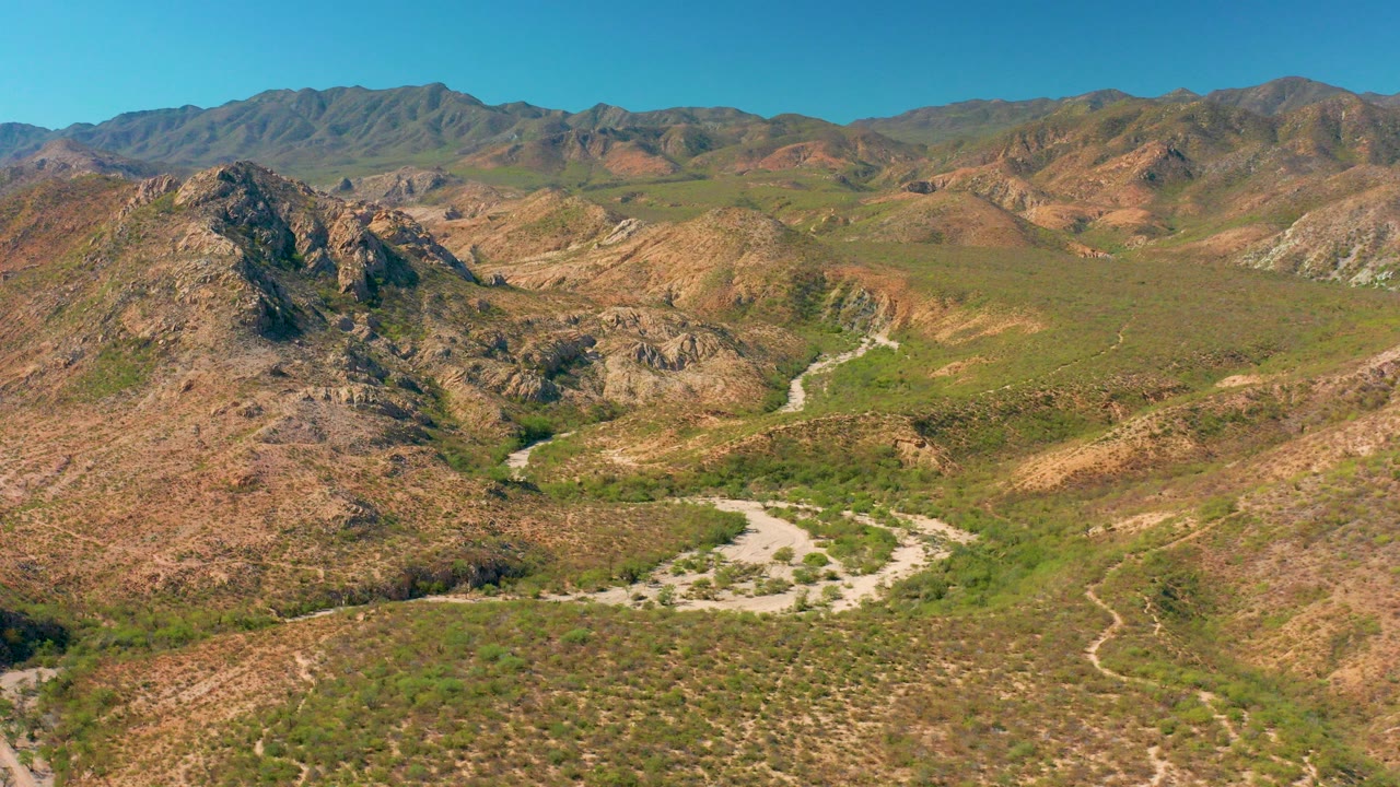 Opening aerial frame of Cabo Pulmo, Baja California Sur, with rocky hills, green-brown desert vegetation, a pale winding dirt path, distant mountains, and no visible people.