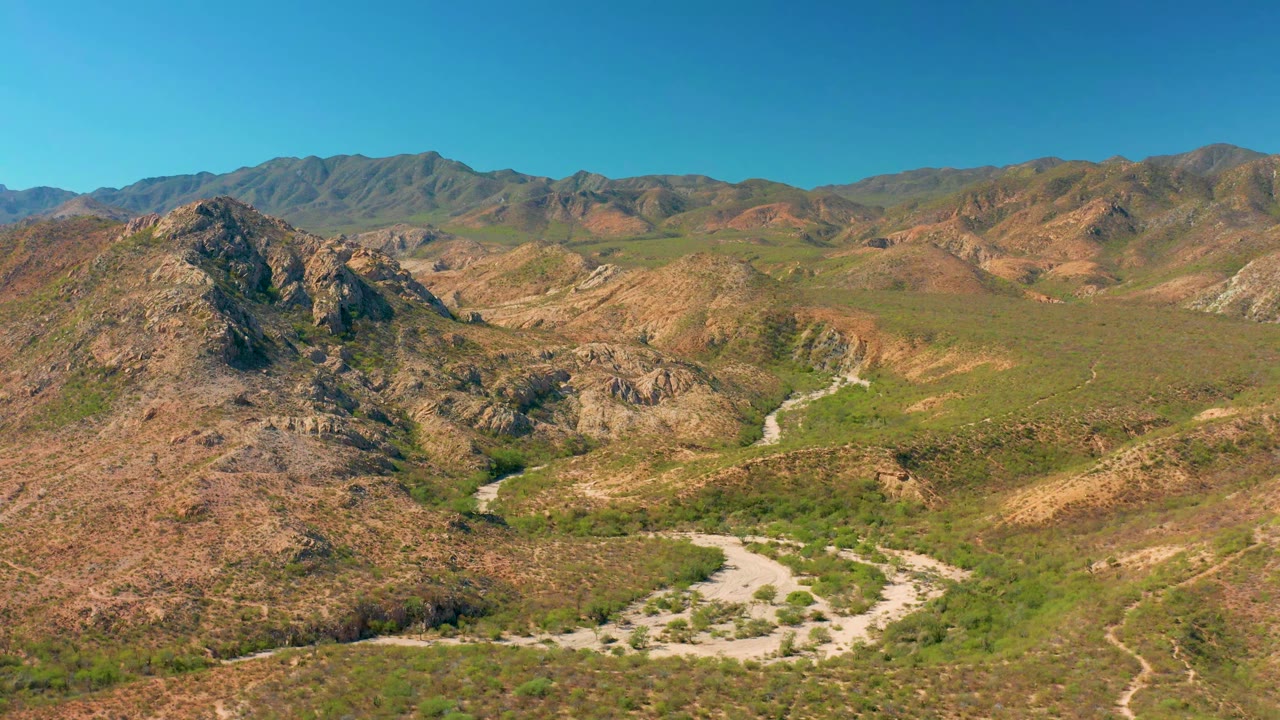 Aerial frame in Cabo Pulmo with a rocky ridge dominating the foreground, a pale dirt road below, distant mountains, clear blue sky, and no visible people.