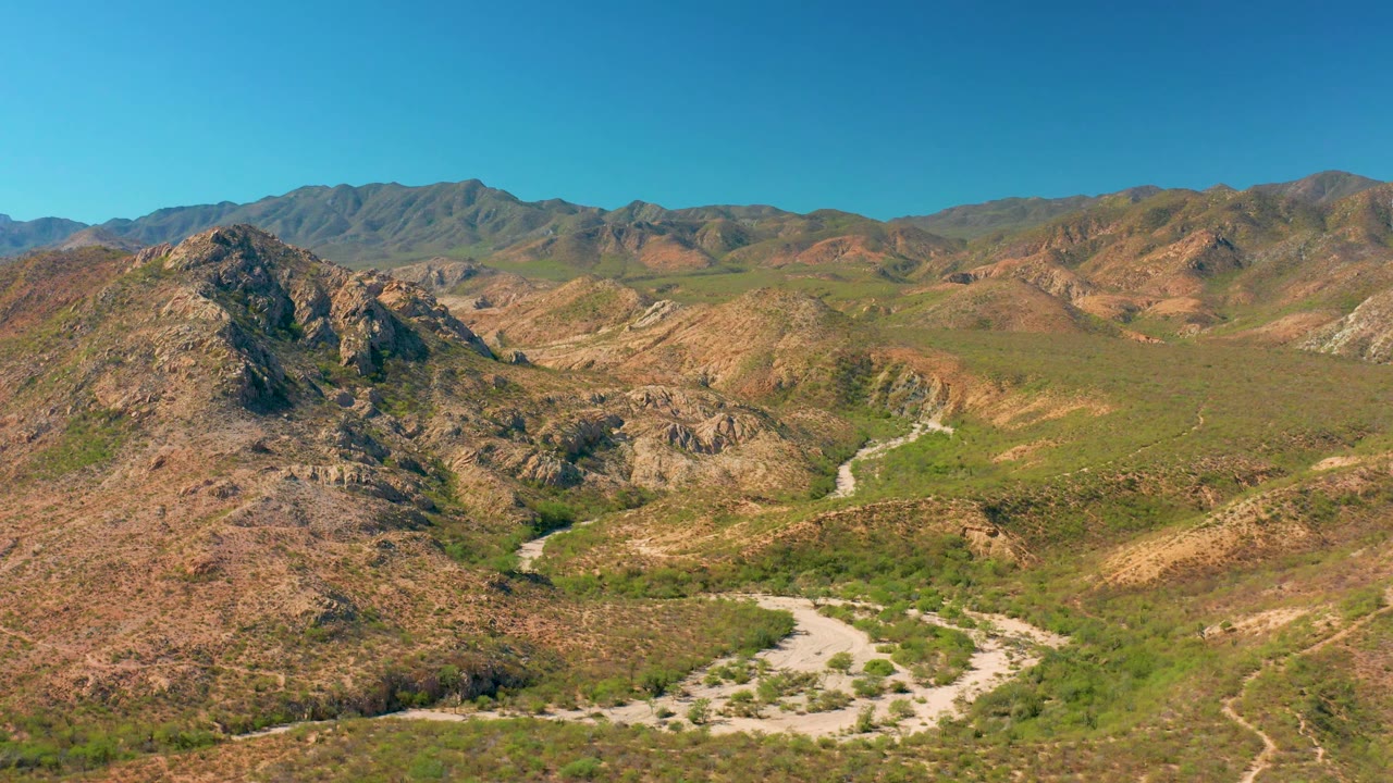 Wide aerial view of Cabo Pulmo terrain where the dirt road bends across the valley floor between scrub-covered hills and layered mountains, with no visible people.