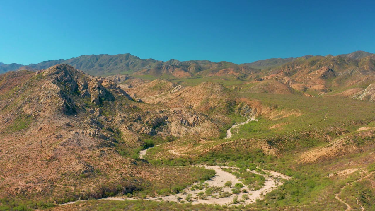 Mid-clip aerial view over Cabo Pulmo, showing the winding dirt road cutting through rocky hills, patchy green vegetation, and brown dry terrain with no visible people.