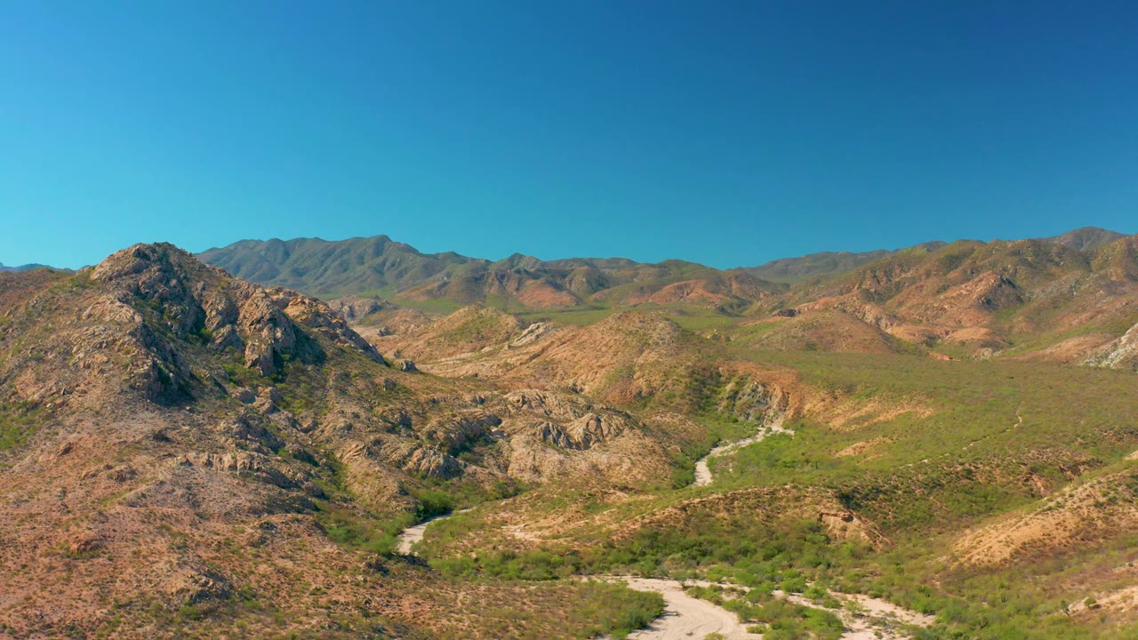 Wide Cabo Pulmo frame showing arid hills on both sides of a green valley floor, a faint winding watercourse, distant mountain ridges, and no visible people.