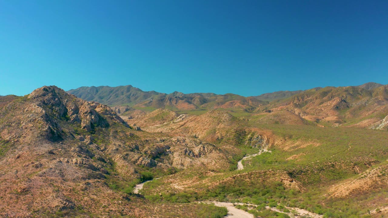 Cabo Pulmo aerial frame with a rocky hill occupying the left foreground, green valley vegetation beyond it, distant mountains, and no visible people.