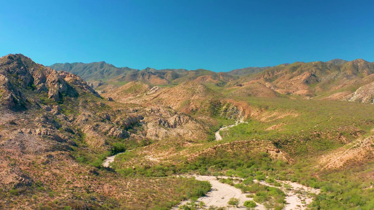 Cabo Pulmo aerial frame showing a rugged hill mass near center, green valley vegetation, a pale track or wash curving through the lower frame, and no visible people.