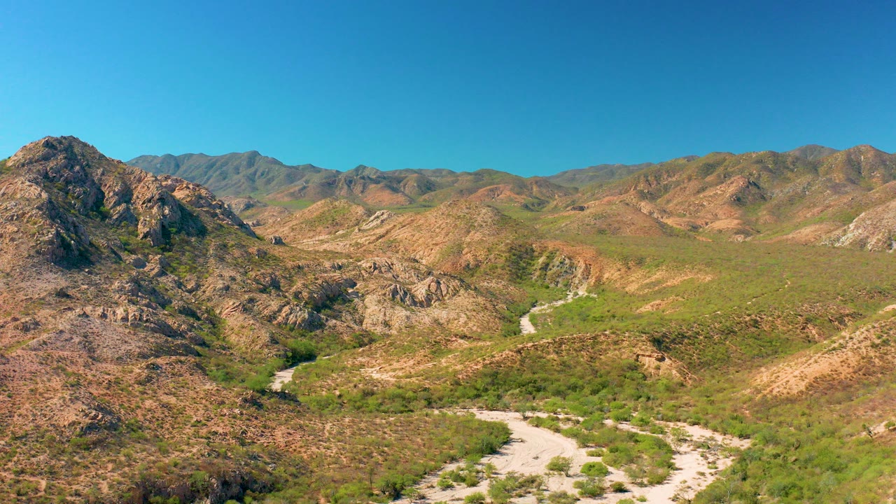 Opening aerial frame over Cabo Pulmo, Baja California Sur, showing a pale dirt road curving through a rocky green-brown mountain valley under a clear blue sky, with no visible people.