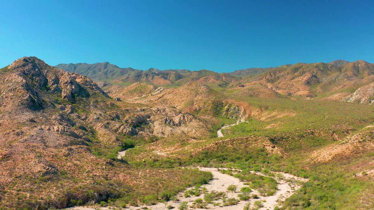Aerial mountain valley with dry ridges and a curving dirt road below.
