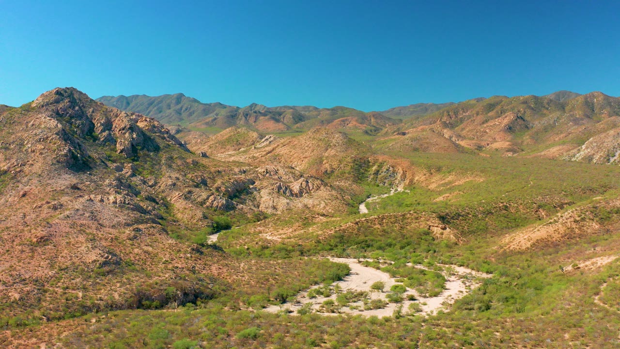 Opening aerial view in Cabo Pulmo, Mexico showing rocky brown hills, green valley vegetation, a pale winding dirt road, distant ridges, and no visible people.