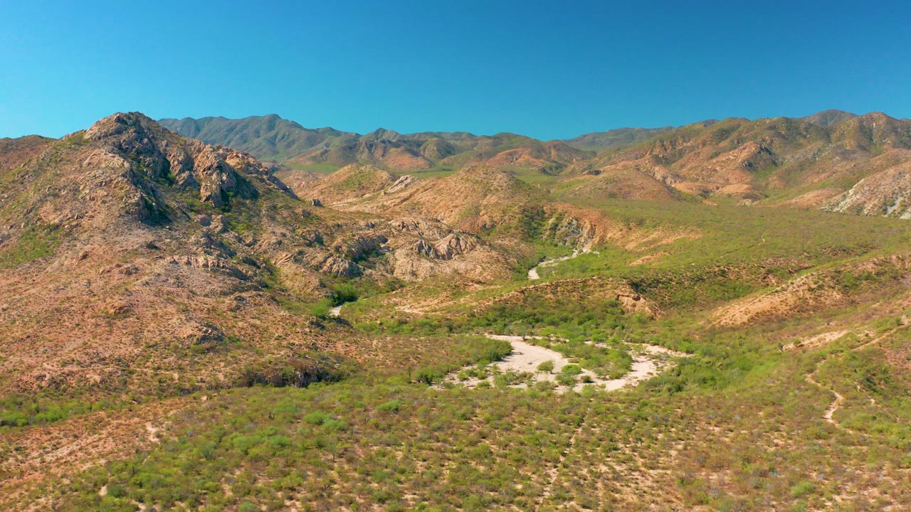 Wide aerial view of rocky desert hills, a pale winding road, green scrub, and a clear blue sky.