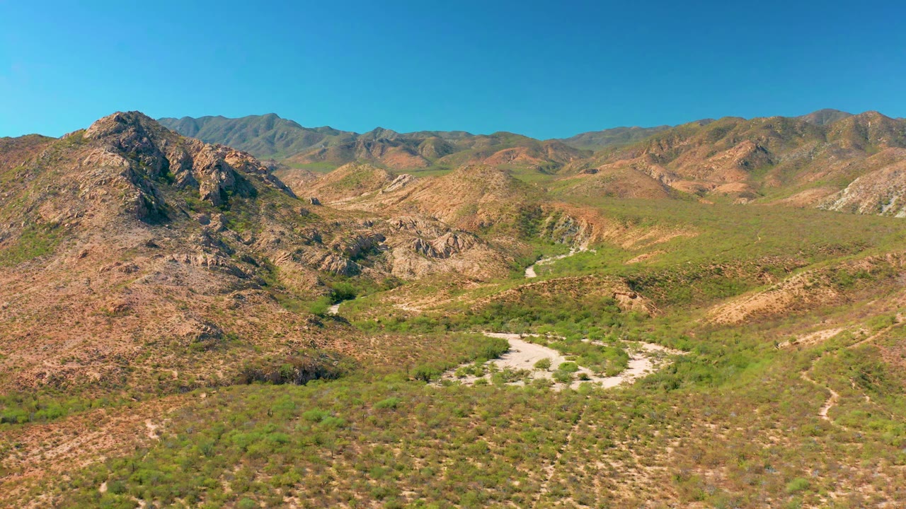 Cabo Pulmo aerial view showing the road and wash slightly more centered between rocky slopes, with green vegetation patches, distant mountains, and no visible people.
