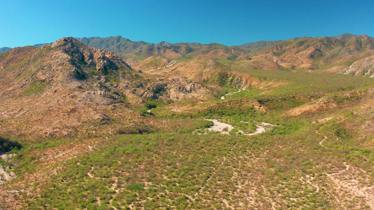 Opening frame over Cabo Pulmo, Baja California Sur, showing a rocky hill at left, sparse green valley vegetation, pale dry washes, distant ridges, clear blue sky, and no visible people.
