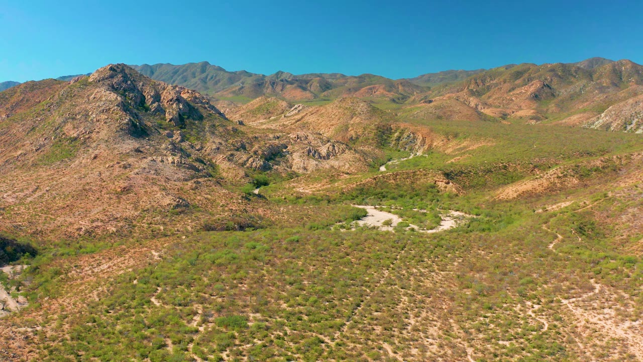 Early frame over Cabo Pulmo with the rocky left slope slightly closer, green-brown valley floor below, dry wash lines through the center, distant mountain ridges, and no visible people.