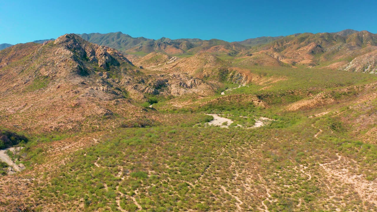 Mid-clip frame over Cabo Pulmo showing the drone farther into the valley, with a rocky slope on the left, low scrub below, pale drainage paths, distant hills, and no visible people.
