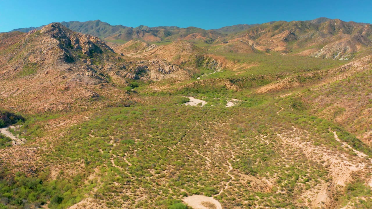 High wide view in Cabo Pulmo showing a central rocky rise, pale dirt road cuts through green-brown scrubland, layered mountain ridges, clear blue sky, and no visible people.