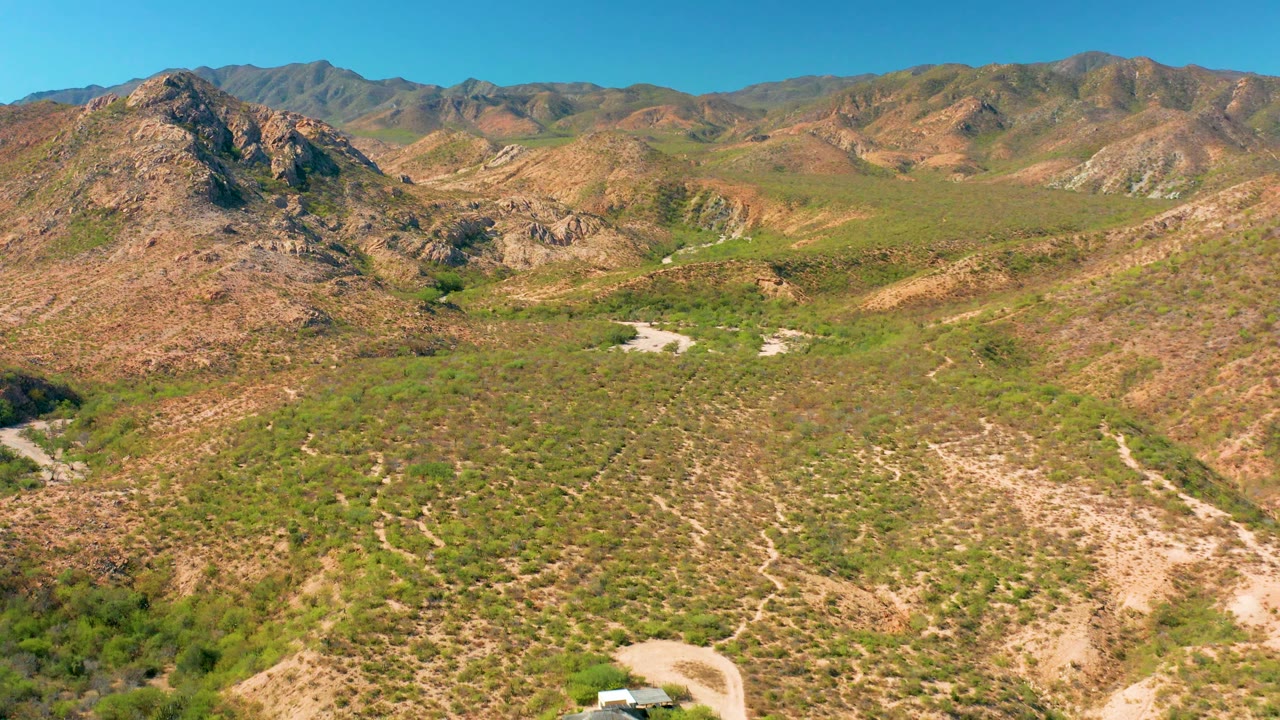 Cabo Pulmo aerial frame with rocky hills, sparse green vegetation, a pale dirt road entering near the lower edge, distant mountains, bright daylight, and no visible people.
