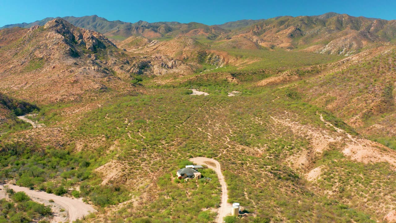 Wide drone view of Cabo Pulmo mountain terrain, with the road entering from the lower edge and rocky ridges crossing the background under clear sky, with no visible people.