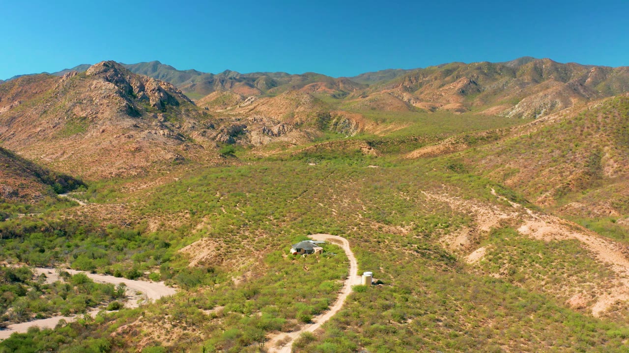 At 0:02, the wide aerial frame shows a vehicle near the winding dirt road, a small building beside the track, dry green-brown hills, and mountains in Cabo Pulmo, with no visible people.