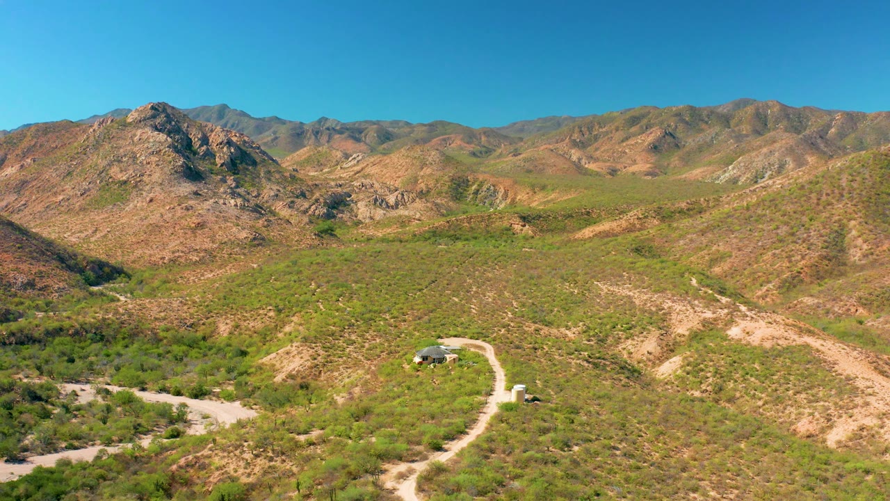 Drone-height view over a dirt road crossing arid hills toward rocky mountains.
