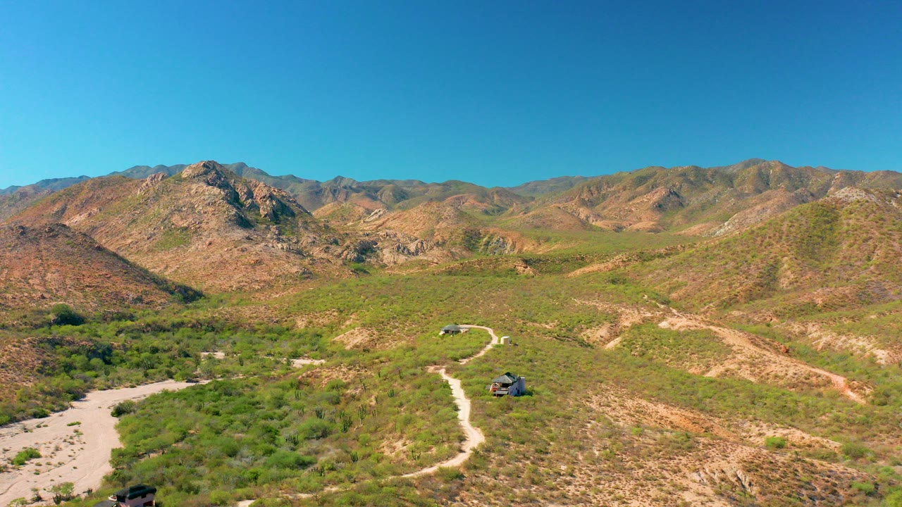 Aerial frame from Cabo Pulmo, Baja California Sur, Mexico showing two small SUVs on a pale winding dirt road below scrub-covered hills and distant mountains, with no visible people.