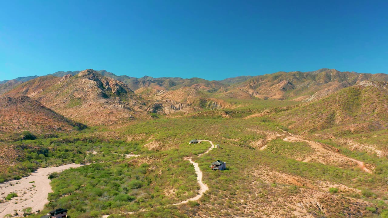 Wide aerial frame in Cabo Pulmo with two off-road vehicles separated along the winding dirt road, scrubland in the foreground, rocky hills behind, and no visible people.