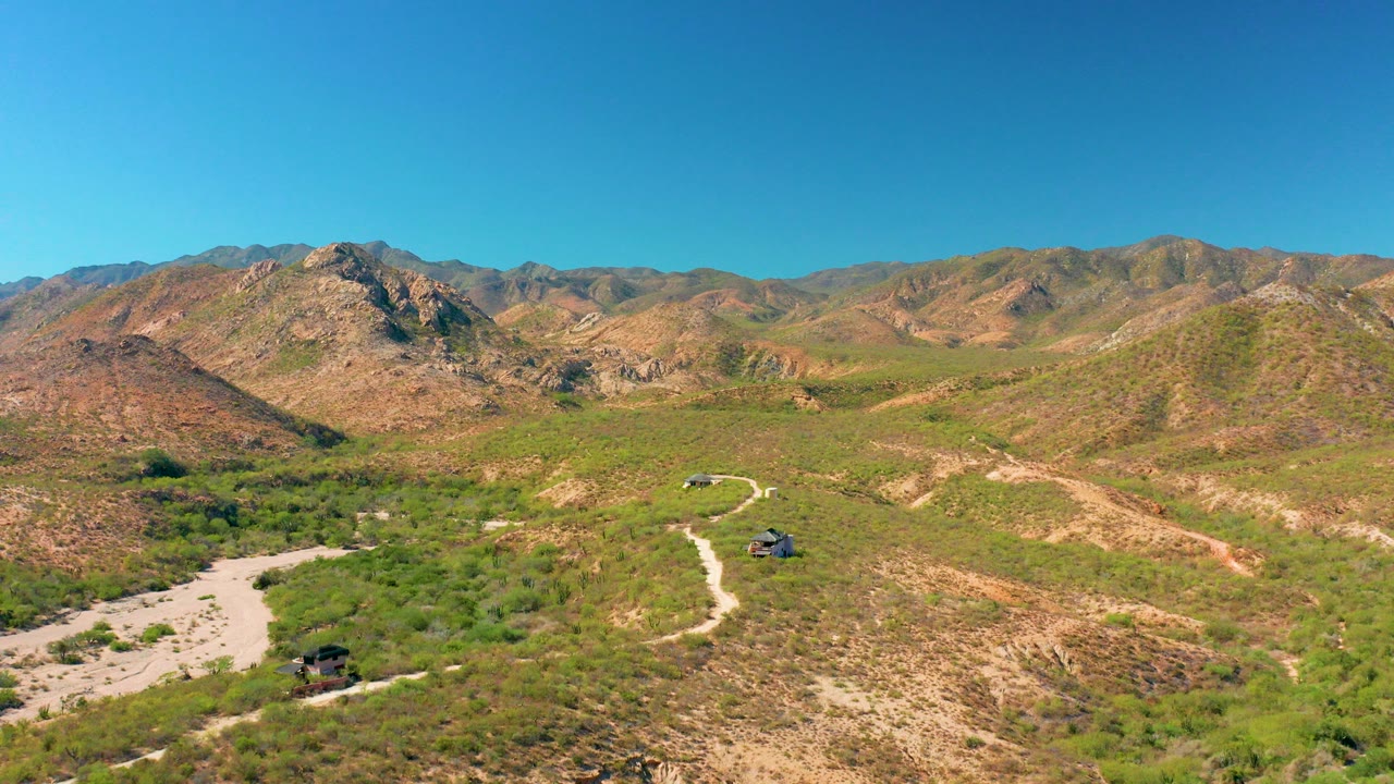 Aerial view over Cabo Pulmo, Baja California Sur, with a pale dirt road winding through scrubby hills, rocky mountains on the horizon, clear blue sky, and no visible people.