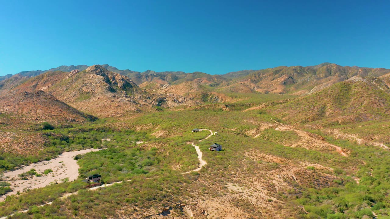 Aerial view of Cabo Pulmo terrain with the dirt road curving through green scrub, two small dark vehicles separated along the route, dry hills, distant mountains, and no visible people.