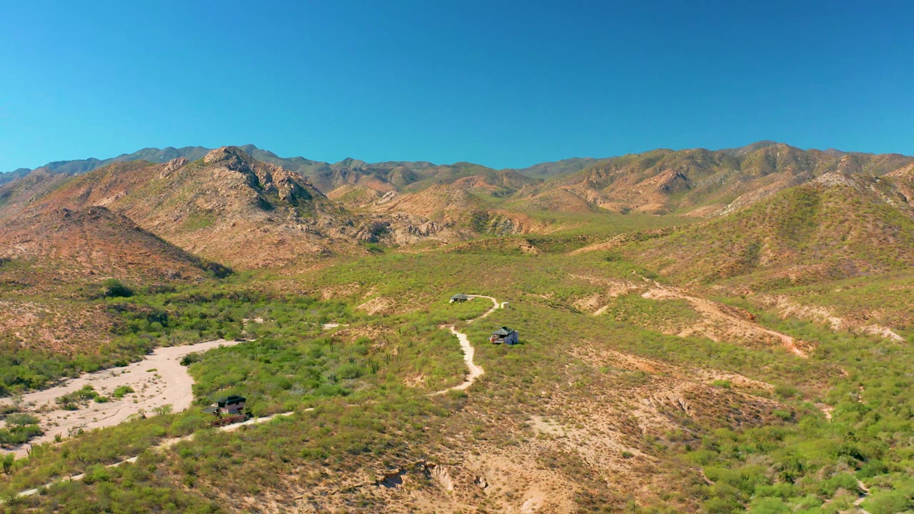 The Cabo Pulmo landscape shows a pale dry wash at left, a winding road through low green vegetation, small vehicles near the road, rugged hills beyond, and no visible people.