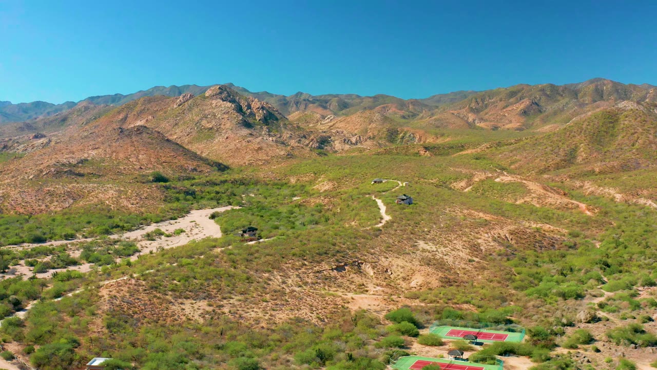 Opening aerial frame over Cabo Pulmo, Baja California Sur, with rocky hills, sparse green scrub, a pale dirt road, distant ridges, and no visible people.