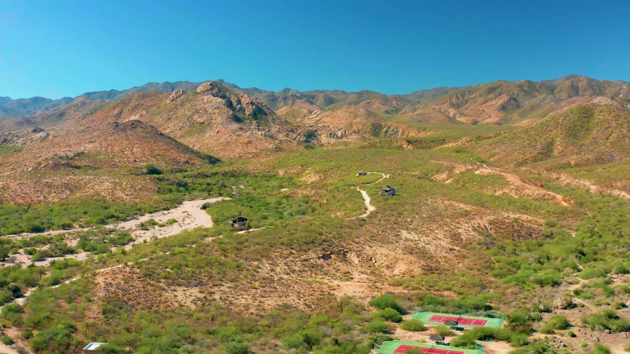 Aerial frame in Cabo Pulmo showing a curving dirt road crossing green desert vegetation below tan rocky slopes, with no visible people.