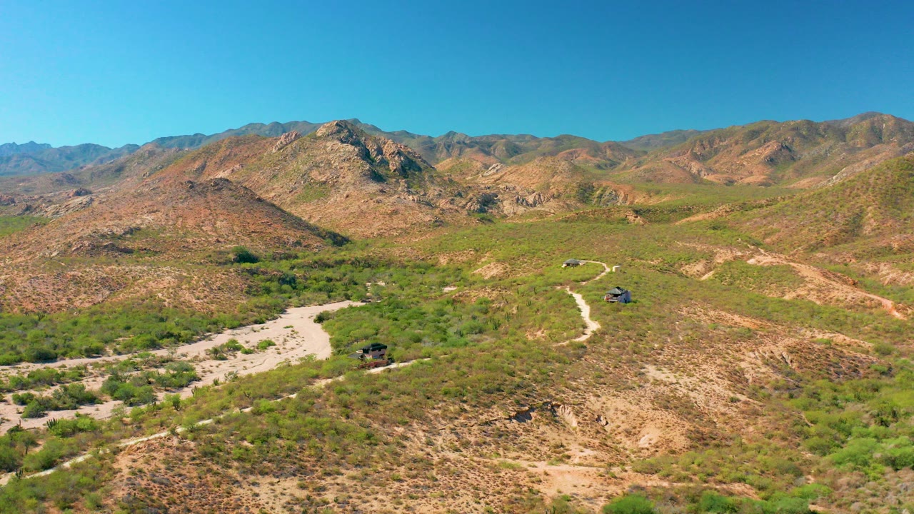 Wide aerial frame over Cabo Pulmo, Baja California Sur, with a pale dirt road curving through scrubby green and brown hills under a clear blue sky, no visible people.