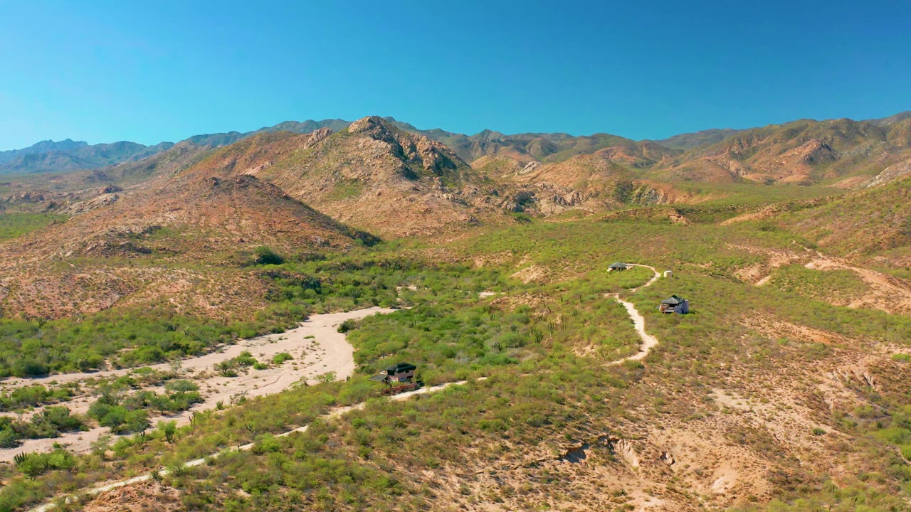 Aerial view in Cabo Pulmo, Baja California Sur, showing two small vehicles separated along a winding dirt road, with tan hills, green brush, rocky ridges, and no visible people.