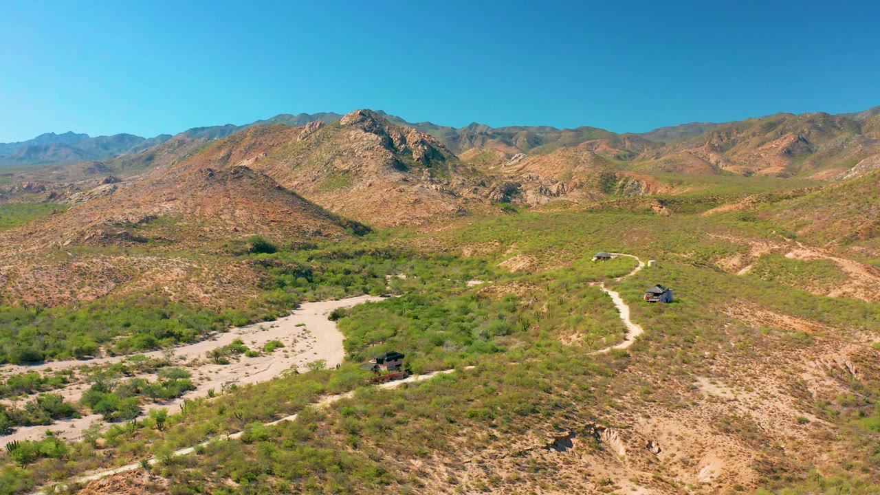 Wide aerial frame in Cabo Pulmo, Baja California Sur, where a pale dirt road bends through green desert vegetation below rocky hills and distant mountains, with no visible people.