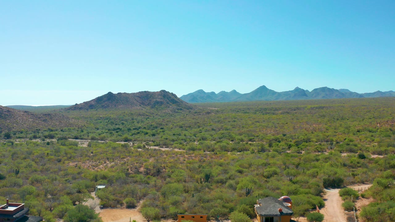 Opening aerial frame over Cabo Pulmo desert scrub with a dirt road and small buildings at the bottom edge, distant blue mountains on the horizon, and no visible people.