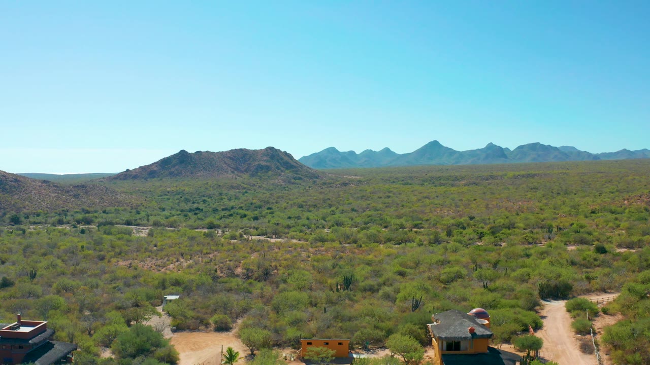 Cabo Pulmo aerial frame with scattered homes near the lower edge, dirt tracks through dense desert scrub, low hills in the midground, distant mountains, and no visible people.