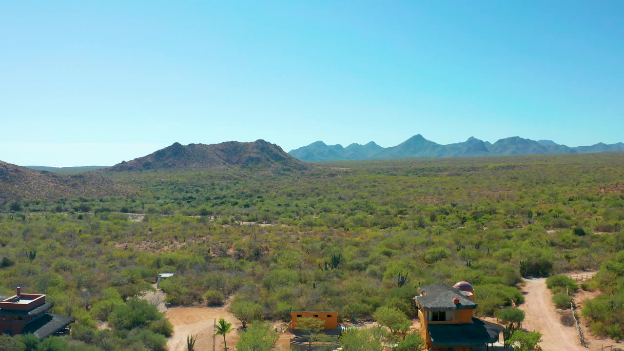 Wide desert aerial with open scrub, a dirt road, small buildings, and clear blue sky.