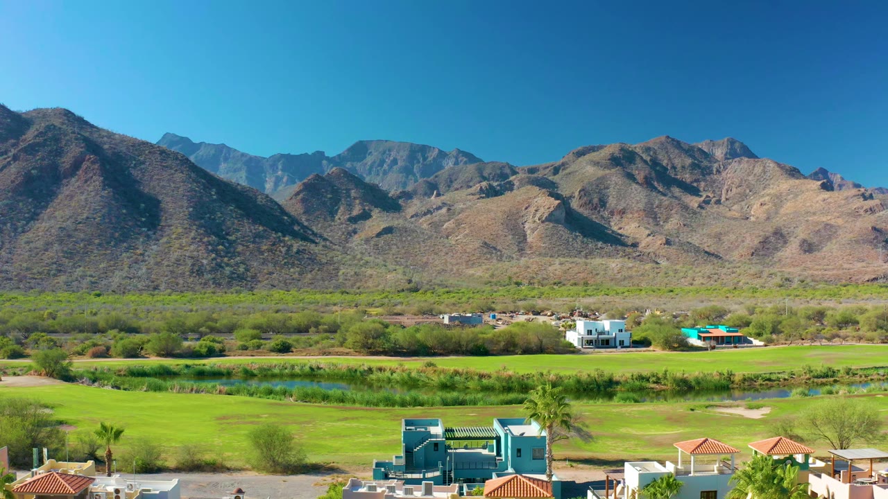 Opening view over Loreto, Baja California Sur with green fields, a narrow water channel, red-roofed homes, a blue building, dry mountains, clear blue sky, and no visible people.