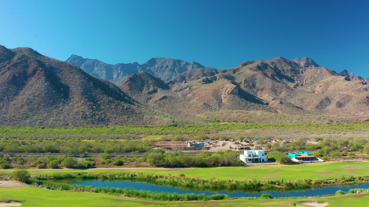 Aerial view in Loreto, Baja California Sur showing blue river water, green fields, small rural buildings, and dry mountain ridges with no visible people.
