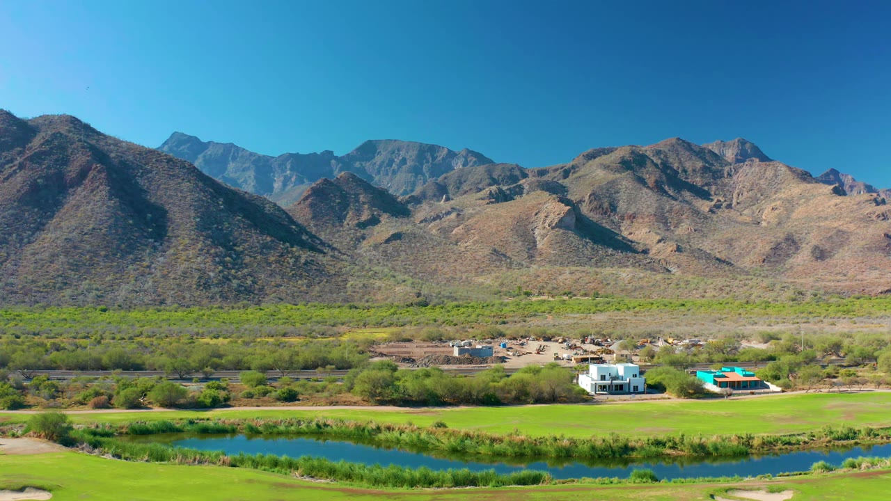 Aerial frame in Loreto, Baja California Sur with the river still visible at the lower edge, bright fields in front, rural buildings at midground, and no visible people.